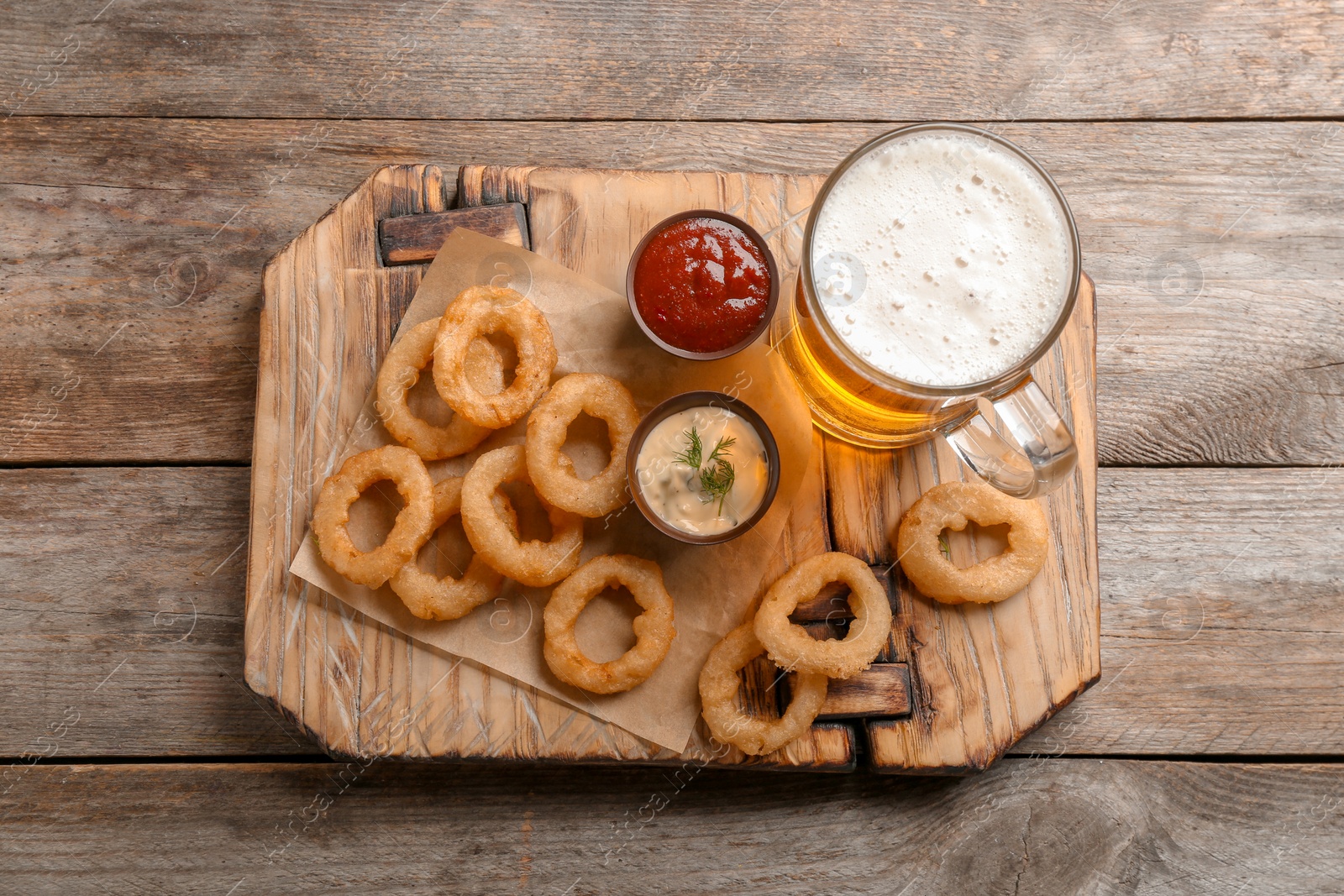 Photo of Fried onion rings served with sauces and beer on wooden board, top view