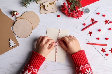 Little child wrapping Christmas card at white wooden table, top view