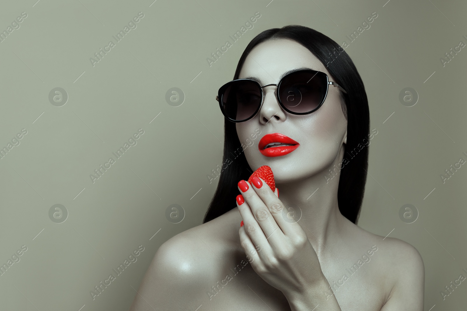 Image of Attractive woman in stylish sunglasses holding strawberry on grey background, space for text. Color accent