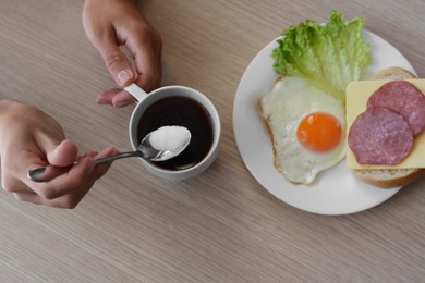 Woman adding sugar to morning coffee while having breakfast at wooden table, top view