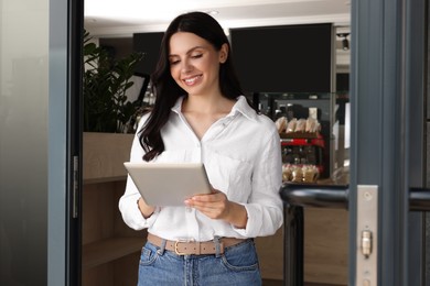 Photo of Happy business owner using tablet at door of her cafe