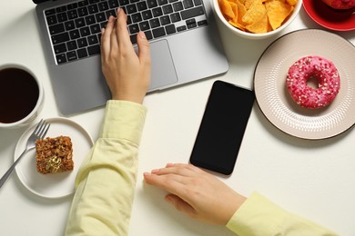 Photo of Bad eating habits. Woman using laptop surrounded by different snacks at white table, top view