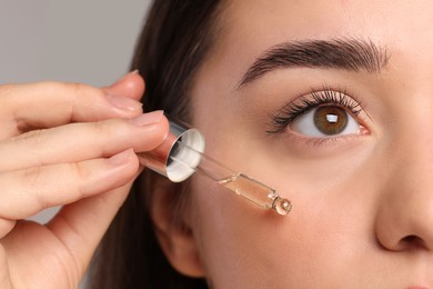 Young woman applying essential oil onto face on light grey background, closeup