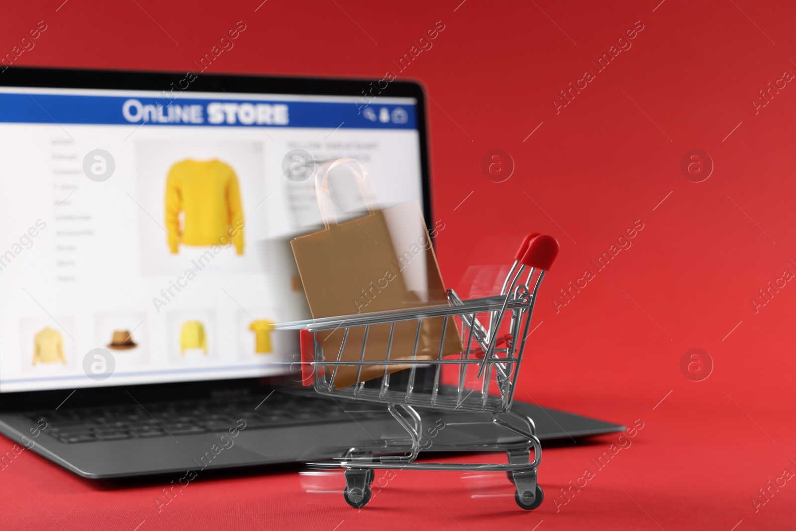 Photo of Shopping bag in mini cart and laptop on red background, selective focus