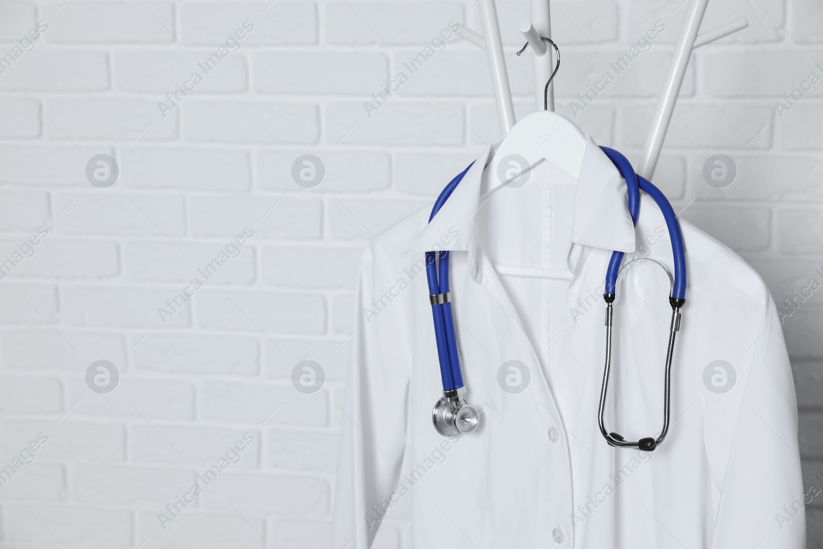 Photo of Medical uniform and stethoscope hanging on rack near white brick wall. Space for text