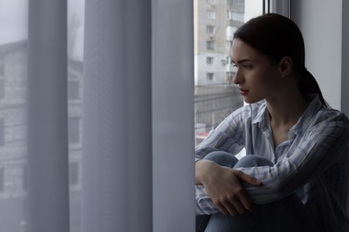 Photo of Melancholic young woman looking out of window indoors, space for text. Loneliness concept