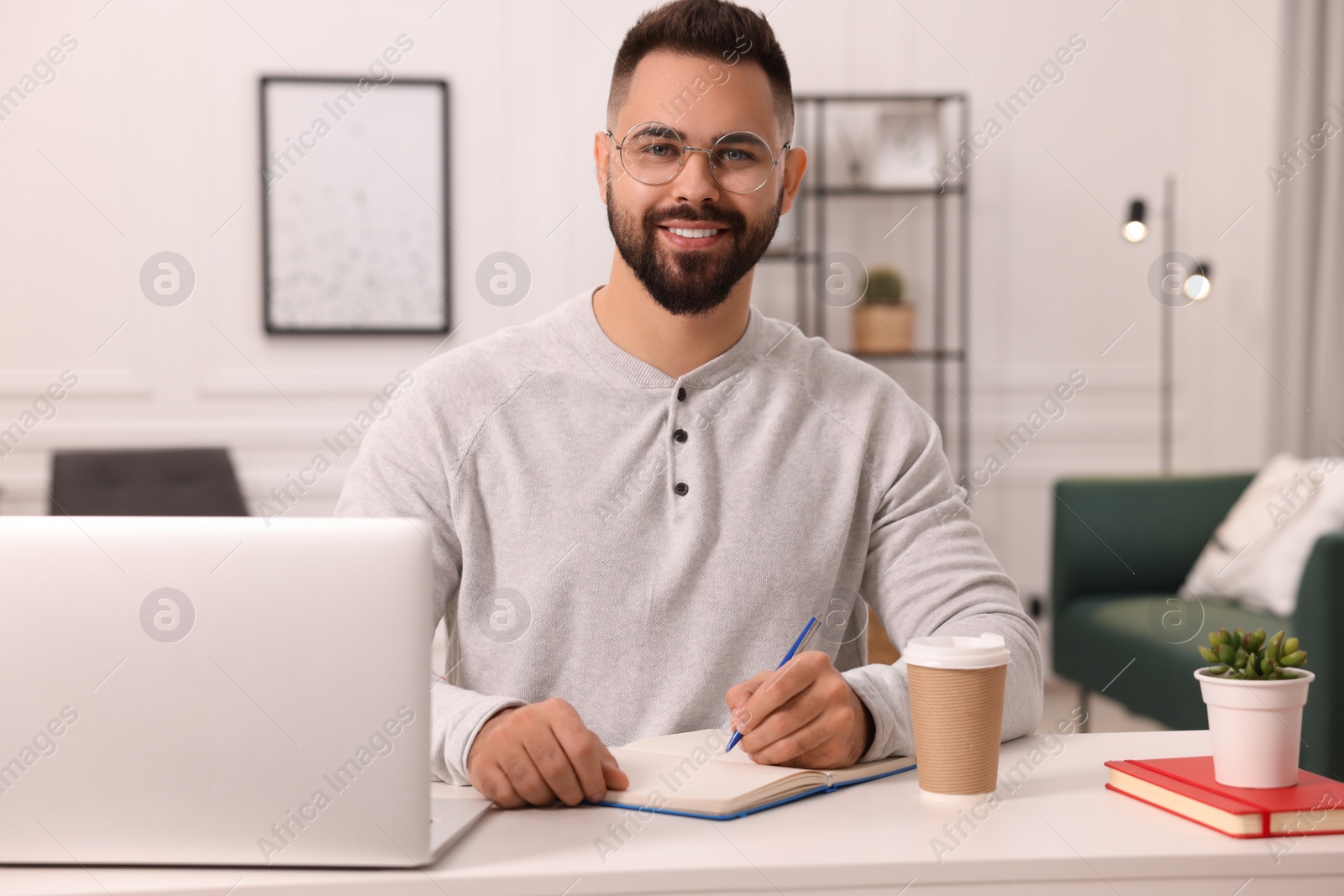 Photo of Young man writing in notebook at white table indoors