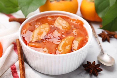 Photo of Bowl of tasty persimmon jam and ingredients on white marble table, closeup