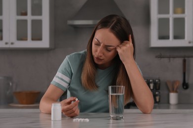 Depressed woman with glass of water and antidepressant pills at table in kitchen