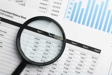 Magnifying glass on accounting documents with data and graph, top view