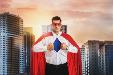 Businessman in superhero cape and mask taking shirt off against beautiful cityscape at sunset 