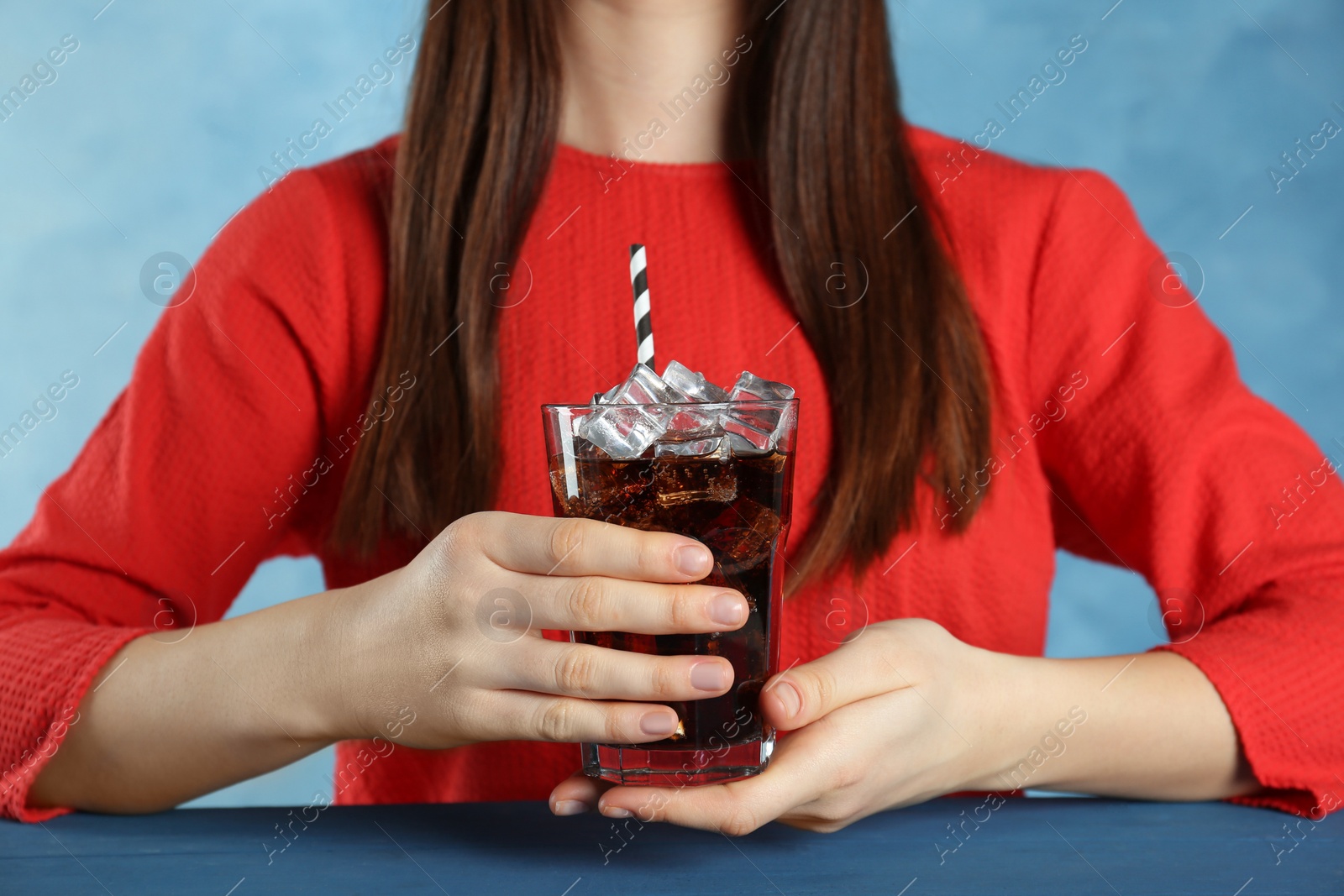 Photo of Woman holding glass of cola with ice and straw at blue table, closeup. Refreshing soda water