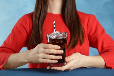 Photo of Woman holding glass of cola with ice and straw at blue table, closeup. Refreshing soda water
