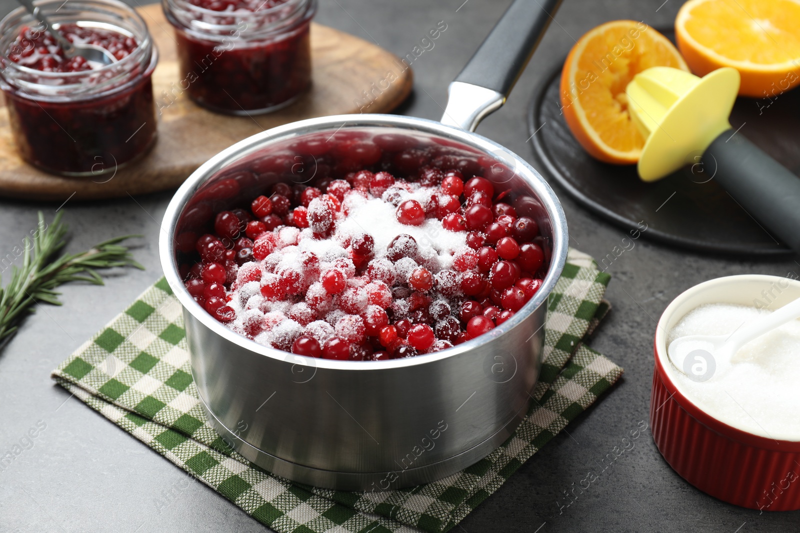 Photo of Making cranberry sauce. Fresh cranberries with sugar in saucepan and ingredients on gray table, closeup
