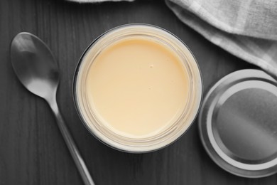 Photo of Open jar with condensed milk and spoon on dark grey wooden table, flat lay