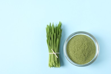 Photo of Wheat grass powder in glass bowl and fresh sprouts on light blue table, flat lay. Space for text