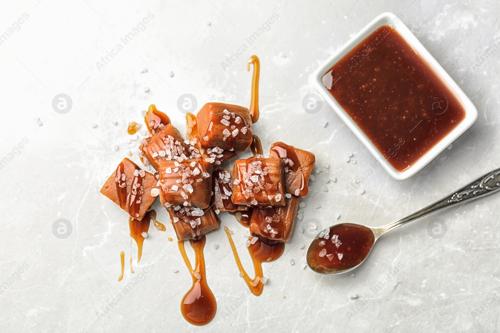 Photo of Spoon, bowl and candies with salted caramel sauce on light background, top view