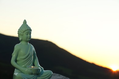 Decorative Buddha statue with burning candle on log in mountains at sunset. Space for text