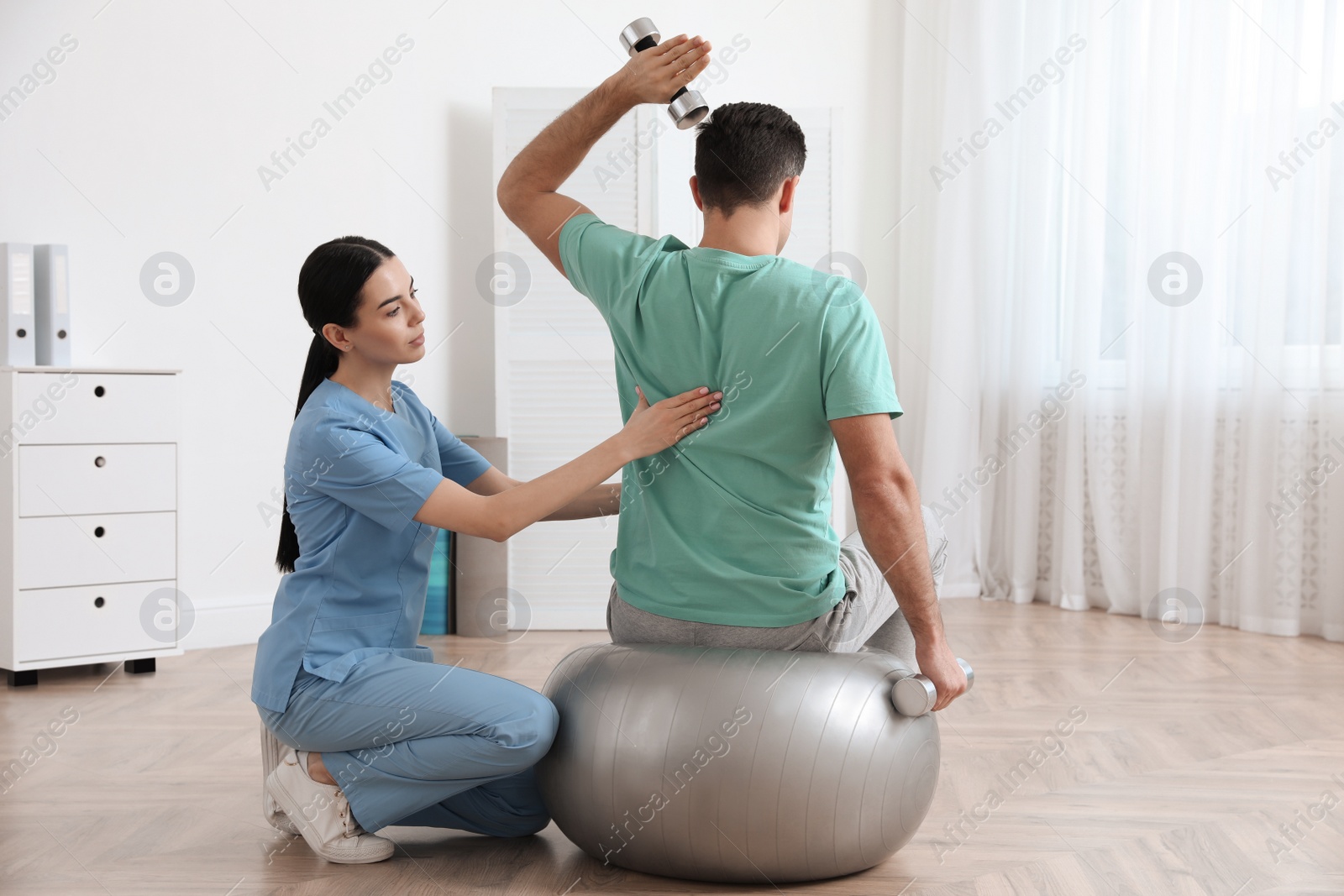 Photo of Orthopedist helping patient to do exercise with dumbbells in clinic. Scoliosis treatment