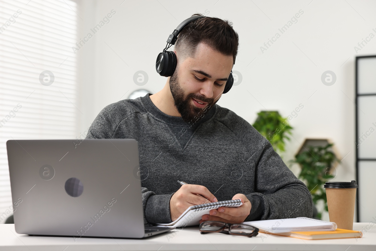 Photo of E-learning. Young man taking notes during online lesson at white table indoors