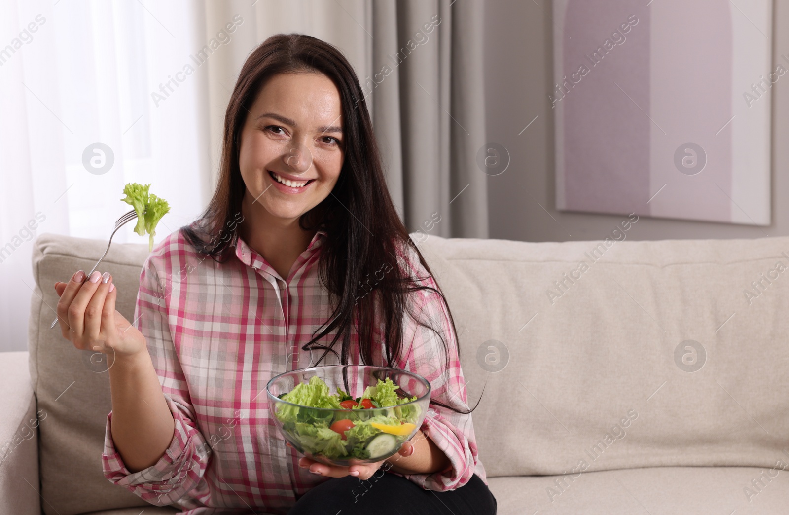 Photo of Beautiful overweight woman eating salad in living room, space for text. Healthy diet