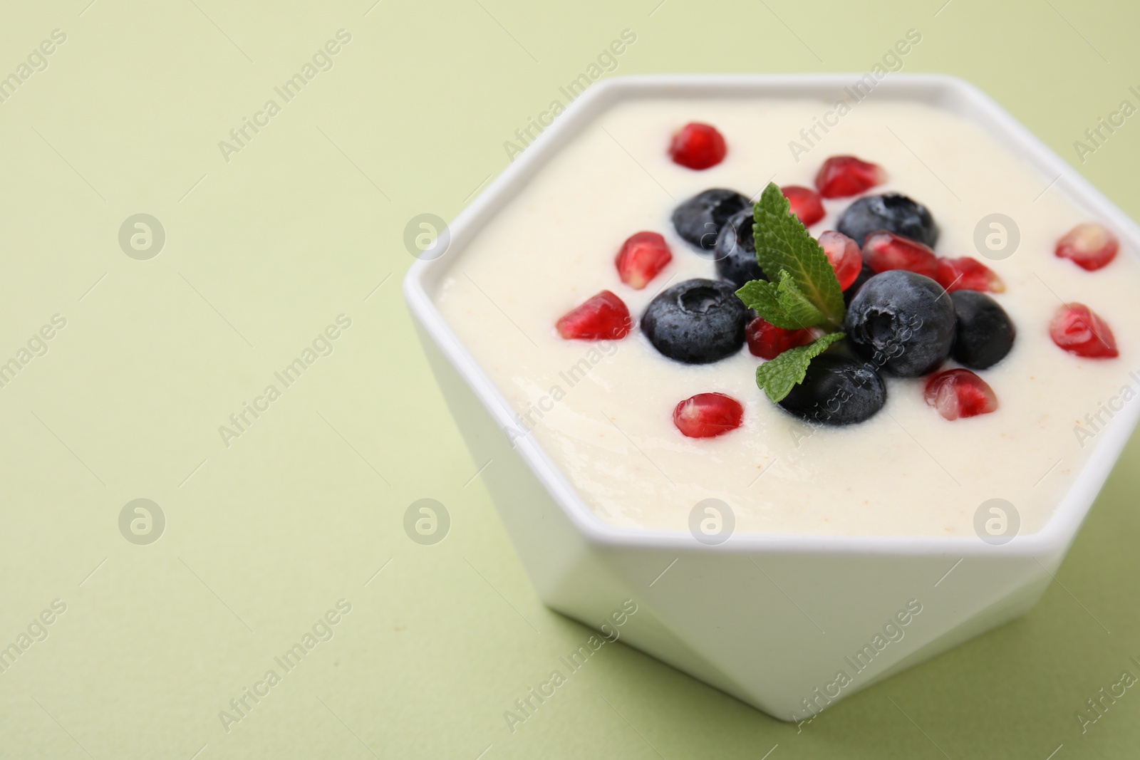 Photo of Bowl of delicious semolina pudding with blueberries, pomegranate and mint on light green background, closeup. Space for text