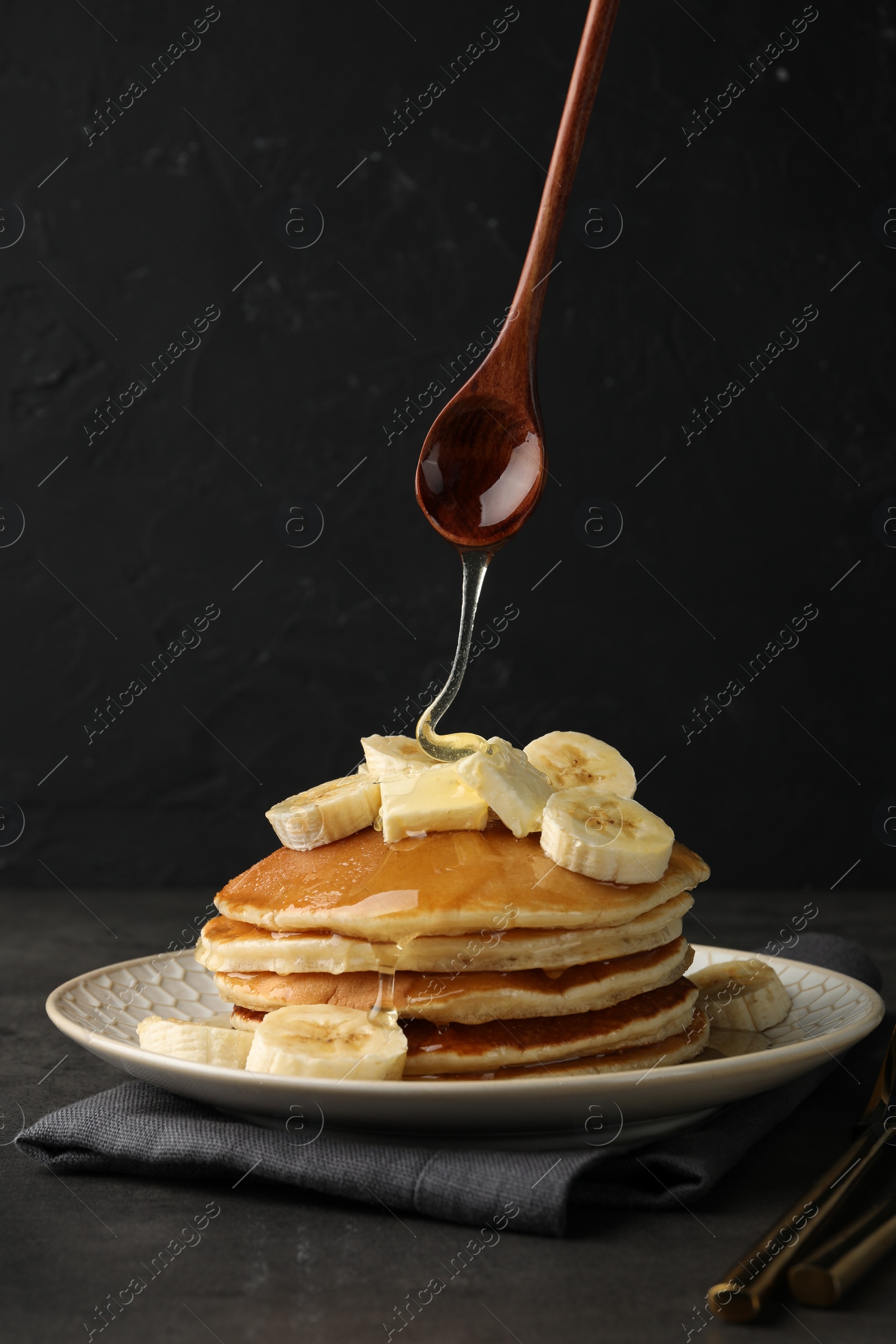 Photo of Pouring honey from spoon onto delicious pancakes with bananas and butter at dark table