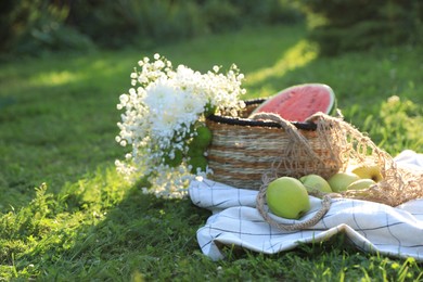 Picnic blanket with tasty fruits, beautiful flowers and basket on green grass outdoors