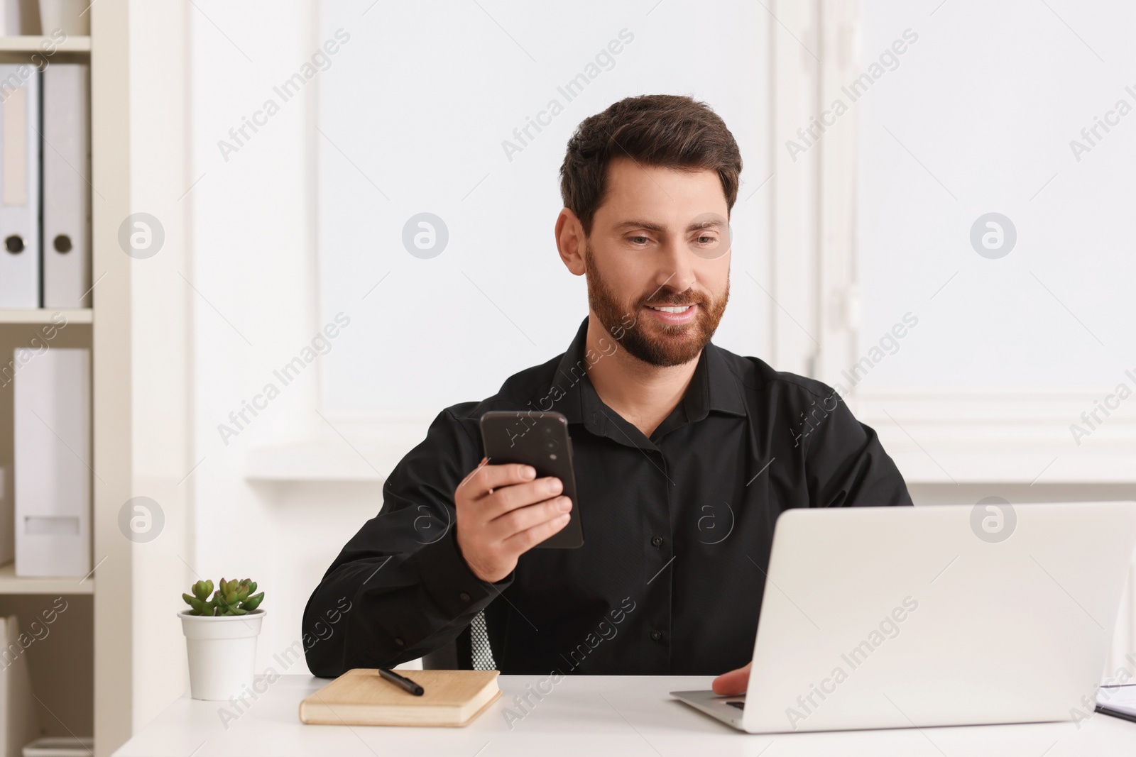 Photo of Smiling man with smartphone using laptop at table in office