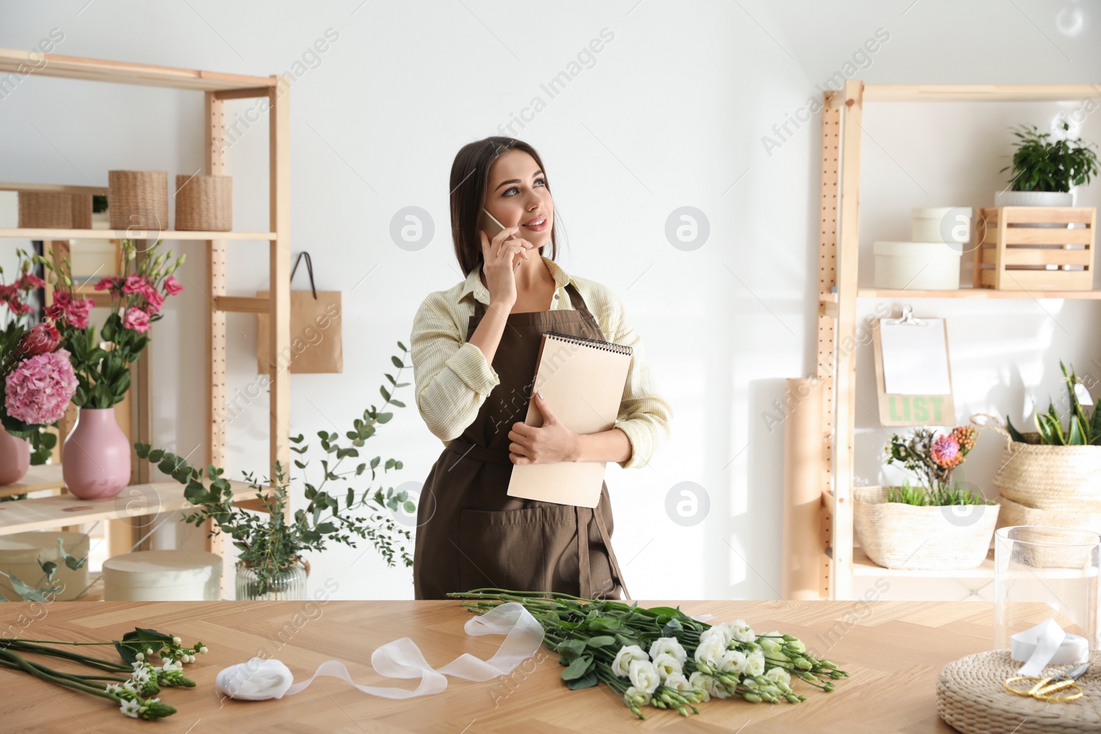 Photo of Florist with notebook talking on smartphone in workshop