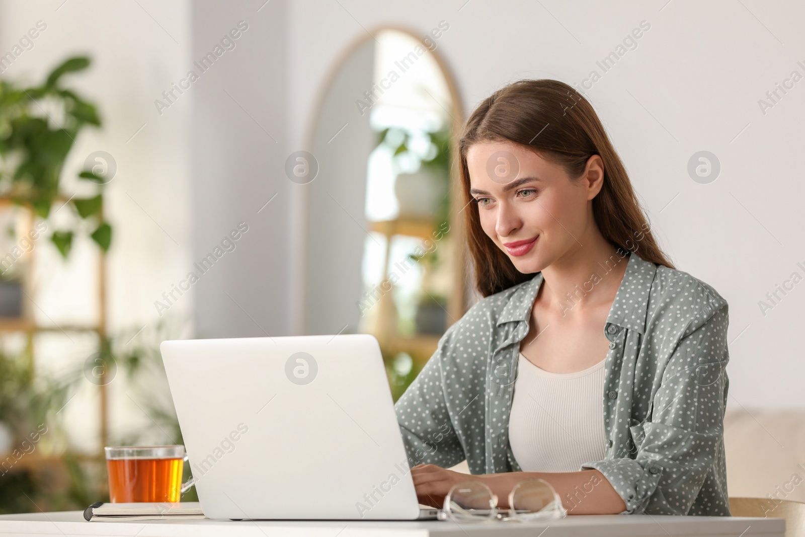 Photo of Happy young woman with laptop and tea at table indoors
