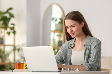 Photo of Happy young woman with laptop and tea at table indoors