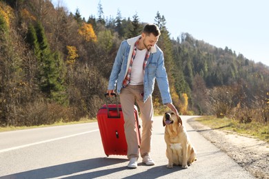 Photo of Happy man with red suitcase and adorable dog on road. Traveling with pet
