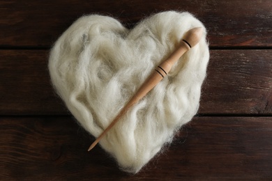 Photo of Heart shaped pile of white wool and spindle on wooden table, top view