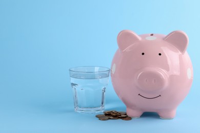 Photo of Water scarcity concept. Piggy bank, glass of drink and coins on light blue background, space for text