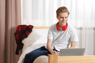 Photo of Online learning. Smiling teenage boy writing in notebook near laptop at home