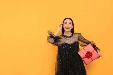 Happy young woman in festive dress with gift box on orange background, space for text