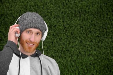 Young man listening to music with headphones near grass wall. Space for text
