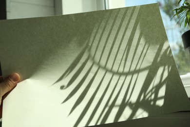 Photo of Woman holding paper sheet with shadow of houseplant near window indoors, closeup