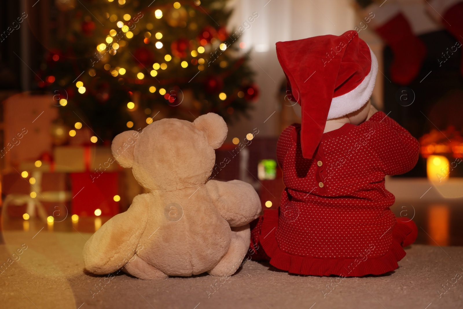 Photo of Baby wearing Santa hat with teddy bear in room decorated for Christmas, back view