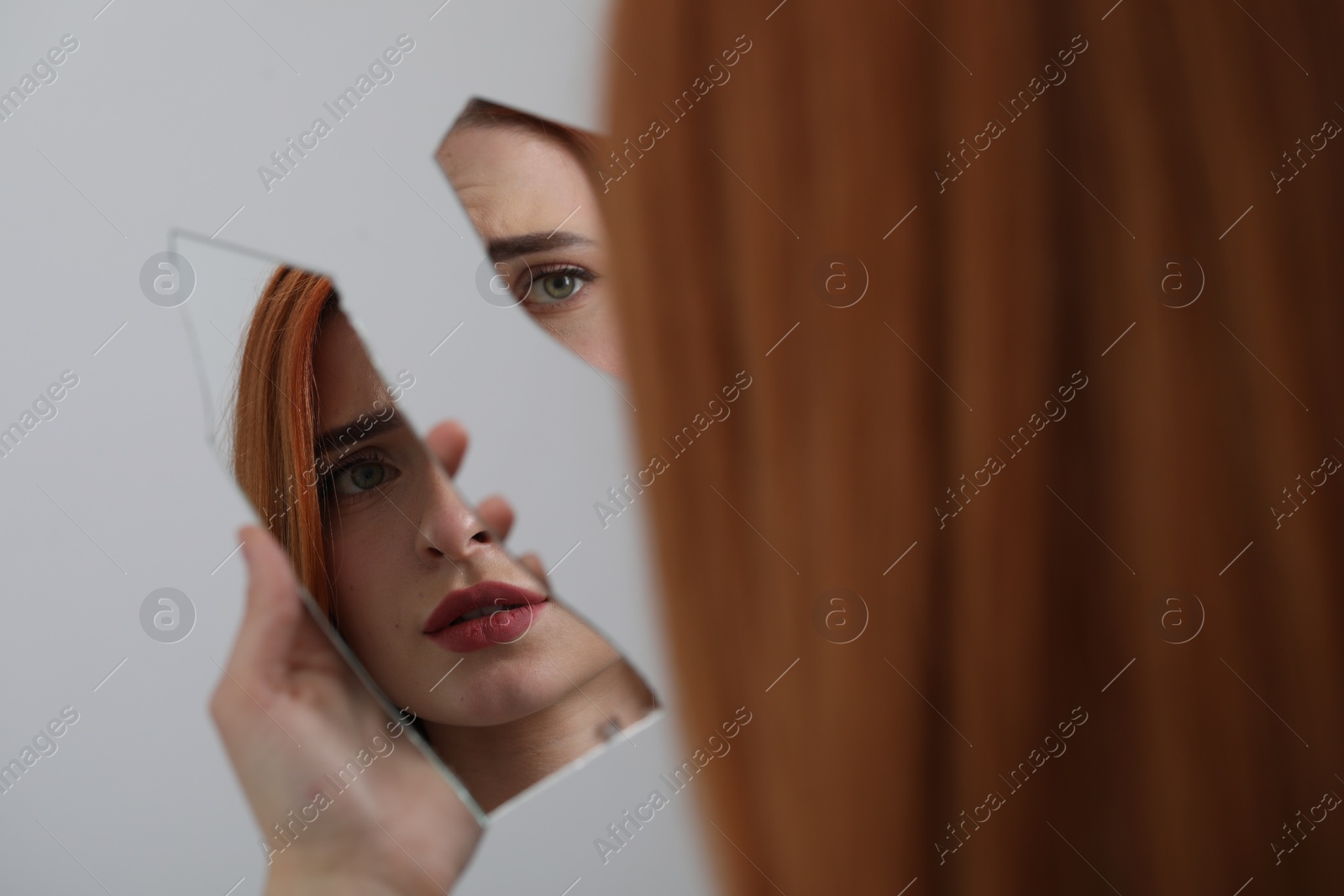 Photo of Young woman looking at herself in shard of broken mirror on light grey background, closeup