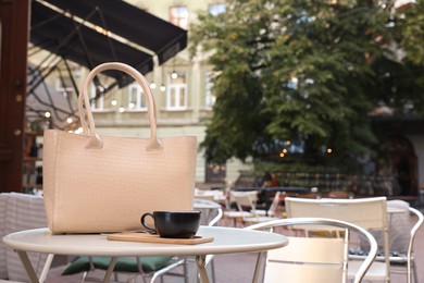 Photo of Stylish bag and cup of coffee on white table in outdoor cafe, space for text