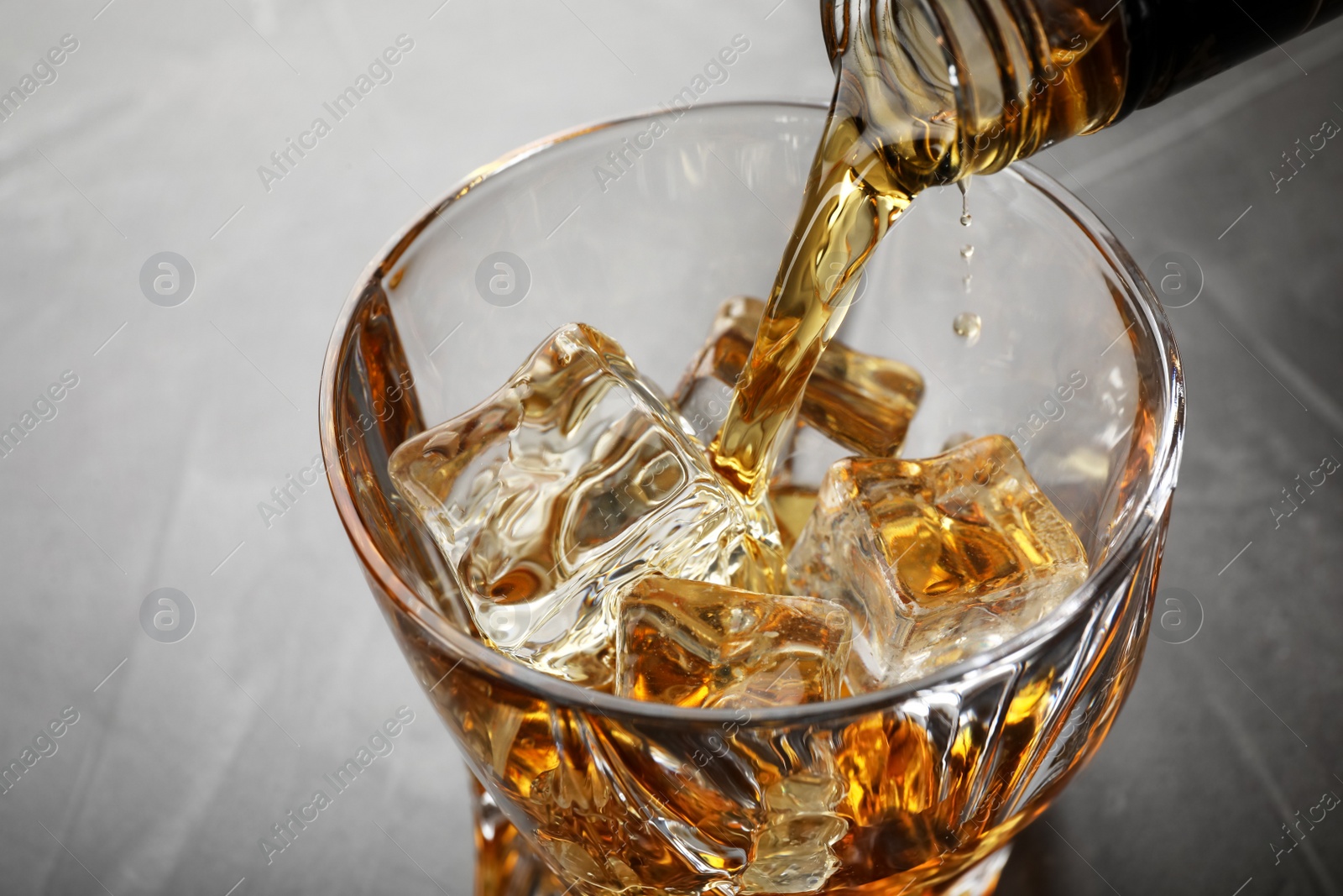 Photo of Pouring whiskey from bottle into glass with ice cubes on table, closeup