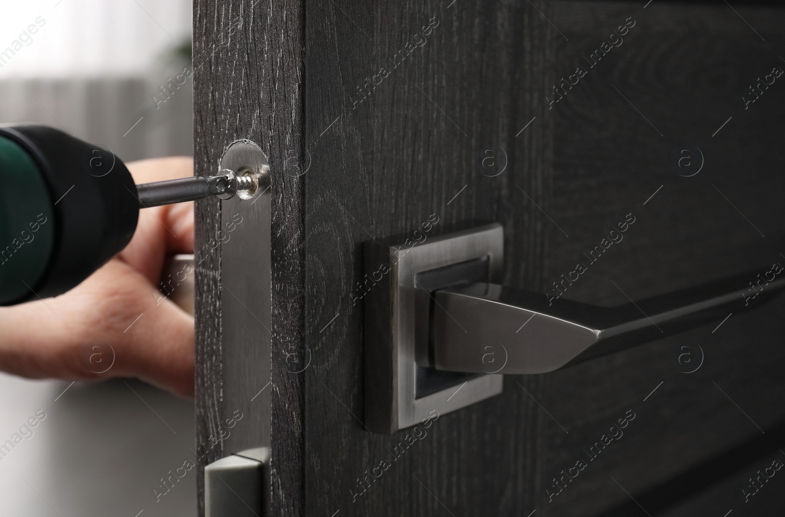 Photo of Handyman with electric screwdriver repairing door handle indoors, closeup