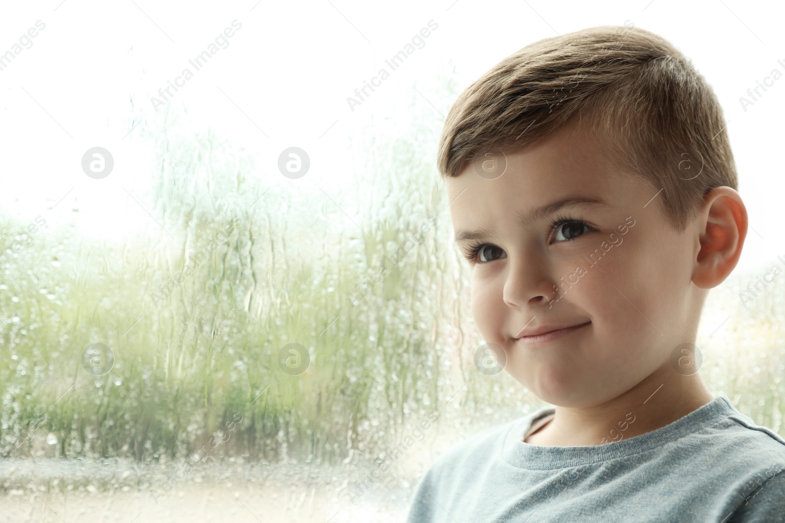 Photo of Cute little boy near window indoors, space for text. Rainy day