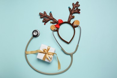 Greeting card for doctor with stethoscope, gift box and reindeer headband on light blue background, flat lay