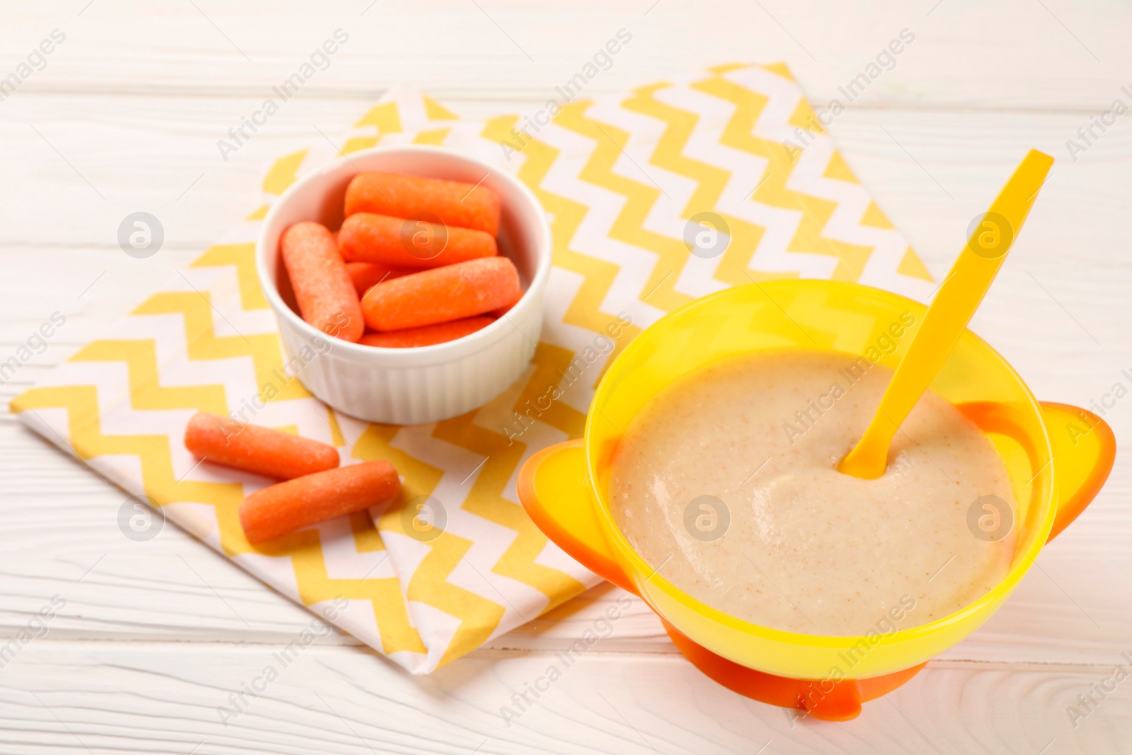 Photo of Baby food. Puree in bowl and small carrots on white wooden table