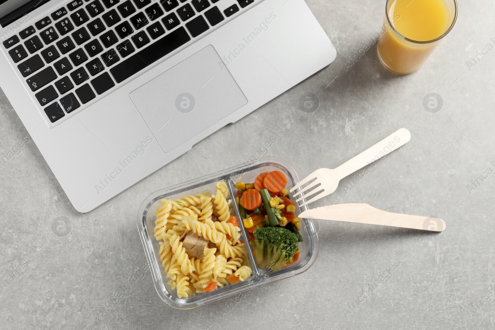 Photo of Container with tasty food, laptop, cutlery and glass of juice on light grey table, flat lay. Business lunch