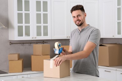Photo of Smiling man taping box with adhesive tape dispenser in kitchen, space for text