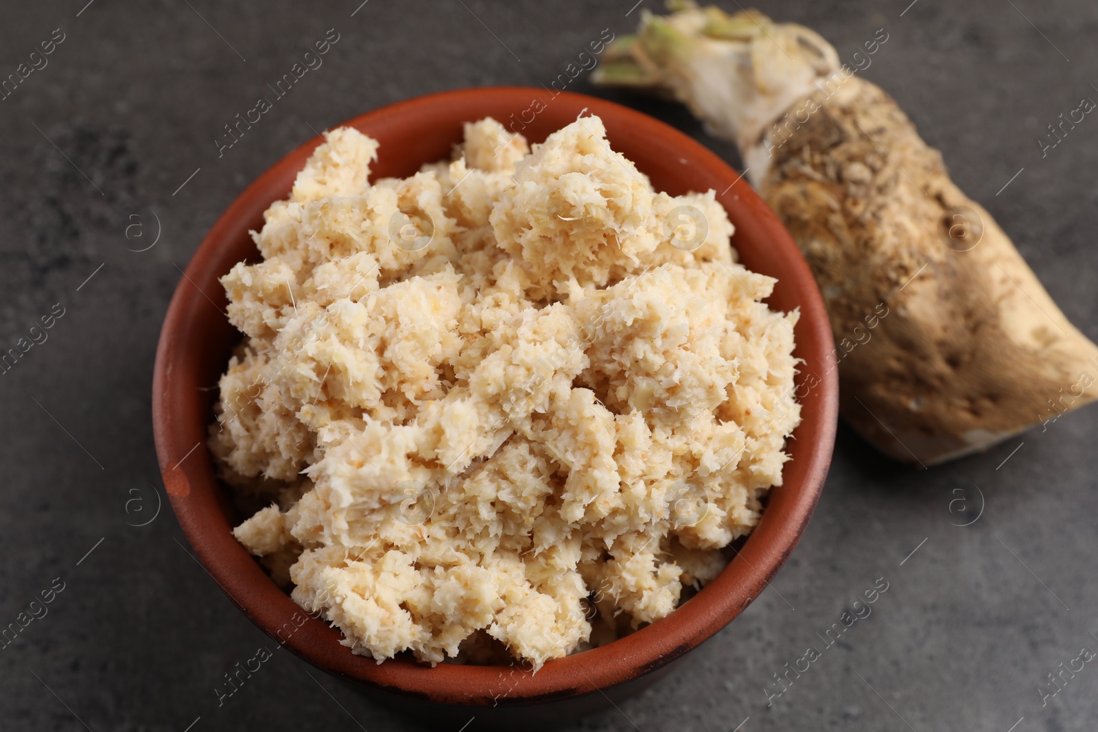 Photo of Bowl of tasty prepared horseradish and root on grey table, closeup